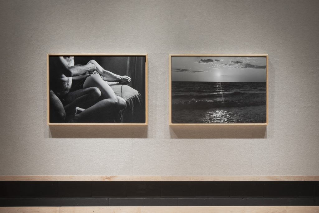 A white wall with two large, rectangular, black and white photographs. The photograph on the left shows a nude man giving an IVF shot to a nude woman laying on a bed. The photograph on the right shows the moon shining across an ocean, with light clouds in the sky.