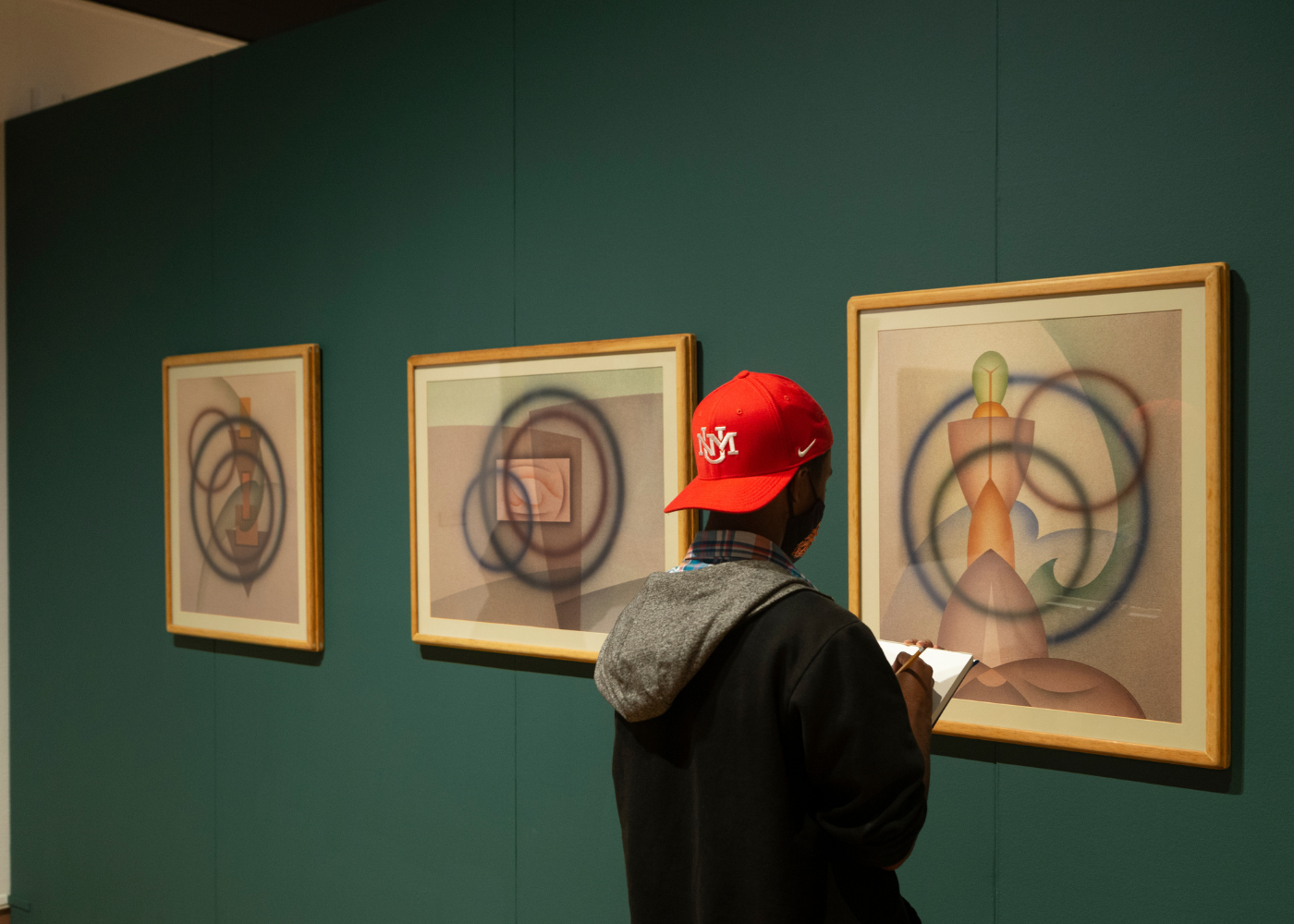 A college student wearing a red UNM hat is photographed while taking notes about a painting in 